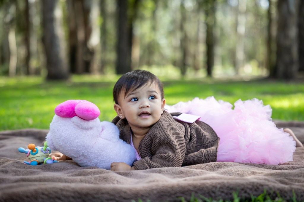 A baby lying on a blanket in a grassy area, wearing a brown sweater and a pink tutu, looking curiously at the camera while holding a stuffed toy.
